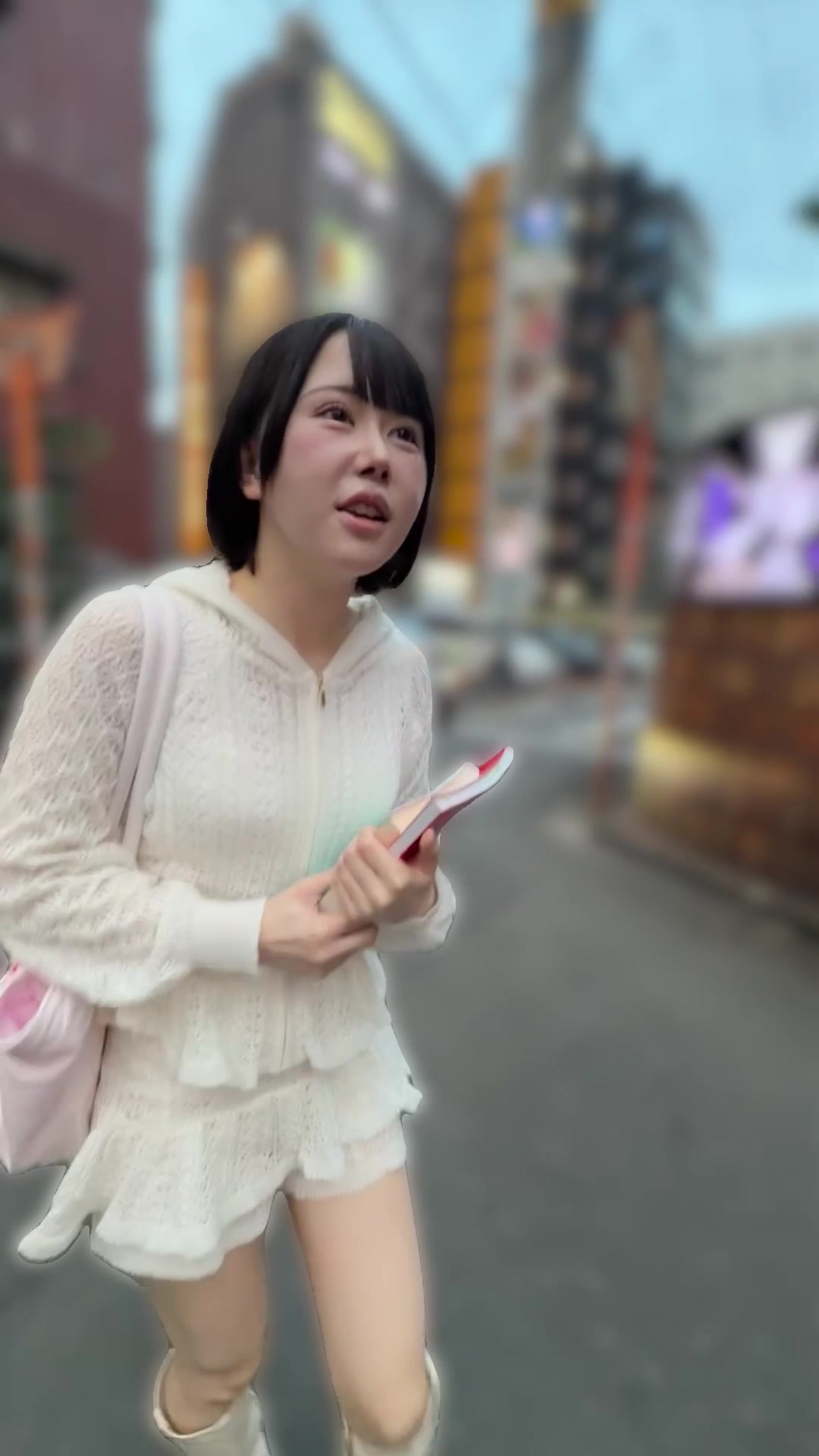 A performer in a white lace hoodie walking down a city street with a pink bag, a clothed still from a video on this page