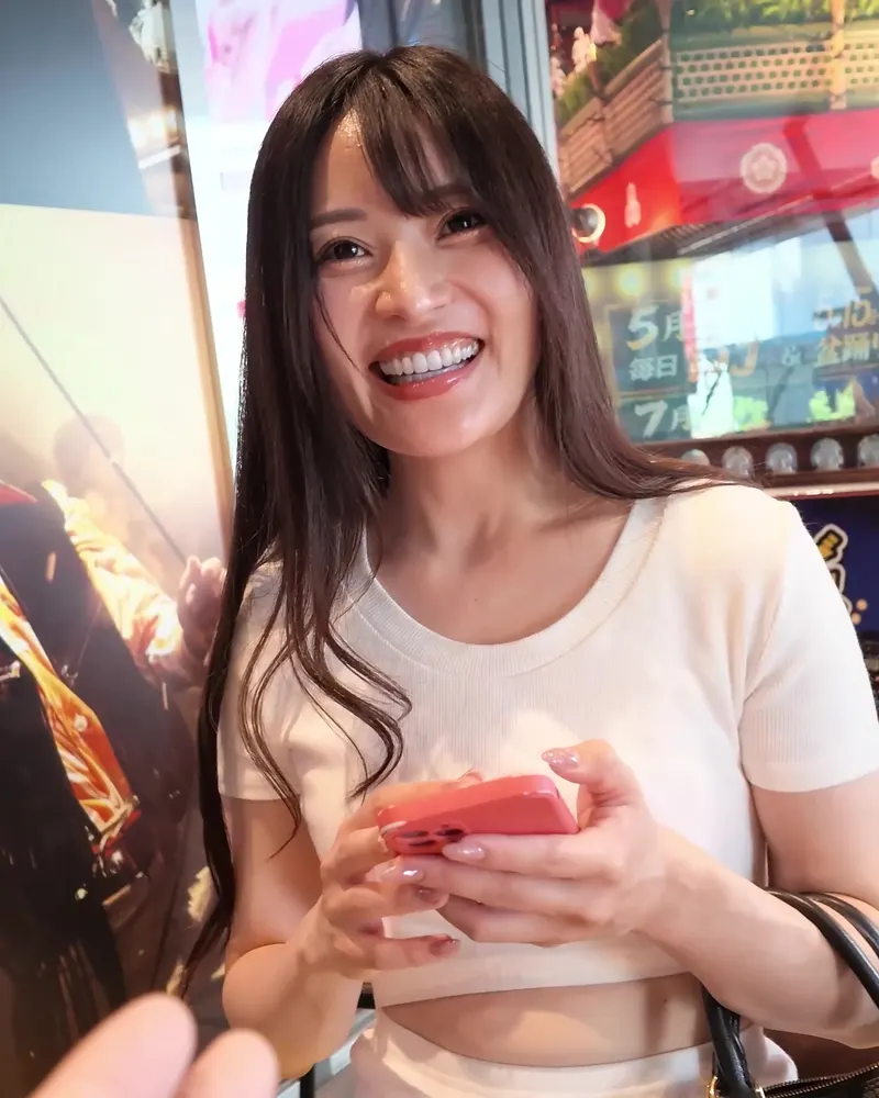 A performer in a white knit top smiling and holding a phone in an entertainment district, a clothed still from a video on this page