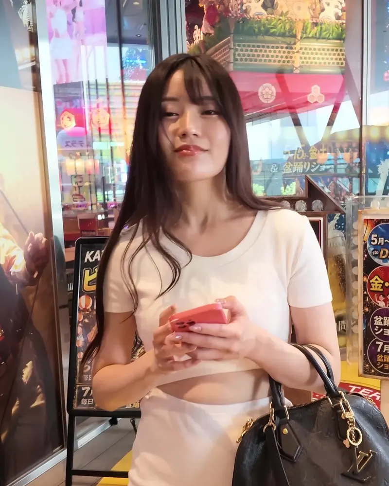 A performer in a white knit top with a black handbag standing by a beer-garden sign, a clothed still from a video on this page