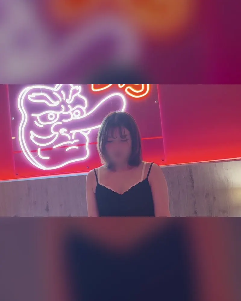 A woman in a black camisole dress in front of a neon sign, a clothed still from a Name-inu video (face blurred in the original)