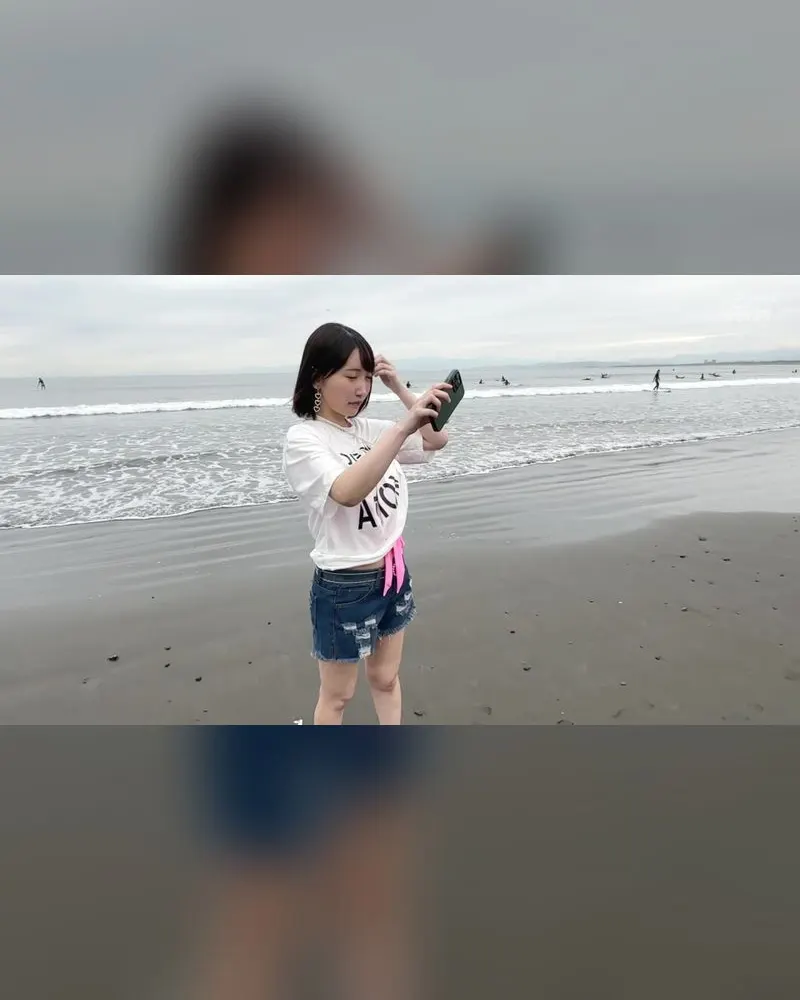 A performer in a white T-shirt and denim shorts taking a photo on a beach, a clothed still from a video on this page
